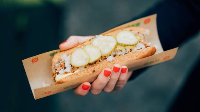 Billedet viser en hånd med rød neglelak, der holder en klassisk dansk hotdog.