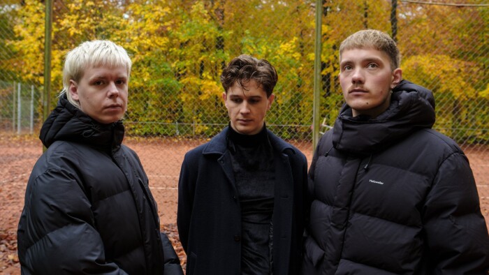 A photograph of the three band members, all wearing black jackets with a background of a metal hatched fence and some green trees.